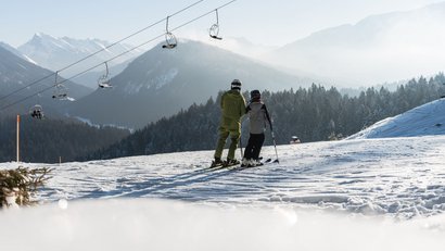 Zwei Skifahrer auf verschneiter Piste mit Berg- und Waldkulisse